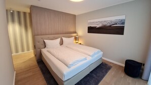 Moderni hotellihuone, jossa on suuri sänky, pääty, yöpöytä lampun kanssa, taulu seinällä ja roskakori.