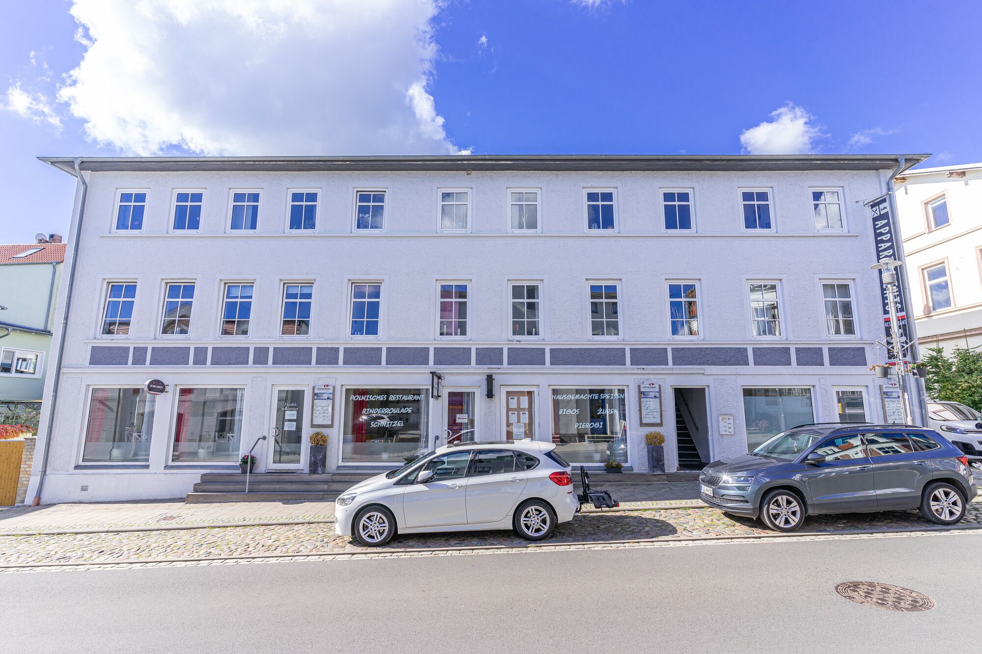 A multi-story building with a row of windows on the ground floor, likely a restaurant or cafe, with cars parked in front.