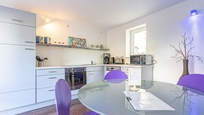 A modern kitchen with white cabinets, a built-in oven, a microwave, and a coffee maker. There is a glass table with two purple chairs and a laptop on it. - Hafenriff - Appartement 1 (Sassnitz)