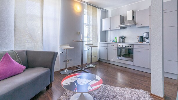 A modern living room with a grey sofa, a small round table with a decorative item, and a kitchen area with grey cabinets and appliances. - Hafenriff - Appartement 4 (Sassnitz)