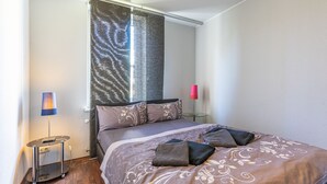 A bedroom with a bed, a small table, a lamp, and a window with a patterned curtain. - Hafenriff - Appartement 4 (Sassnitz)
