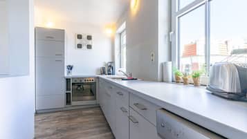 Eine kompakte Küche mit Einbauofen, Kaffeemaschine und Toaster. Auf der Fensterbank stehen Blumentöpfe und das Fenster bietet einen Blick nach außen.