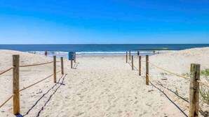 Une plage de sable bordée d’une clôture en bois qui mène à l’océan, un ciel dégagé et quelques personnes visibles au loin.