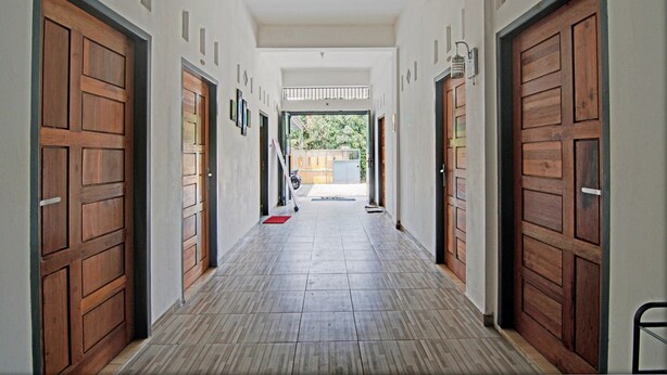 A corridor with wooden doors, a tiled floor, and a view of a garden through an open door at the end. - Hotel O Penginapan Budhe Sri (Koealabadak)