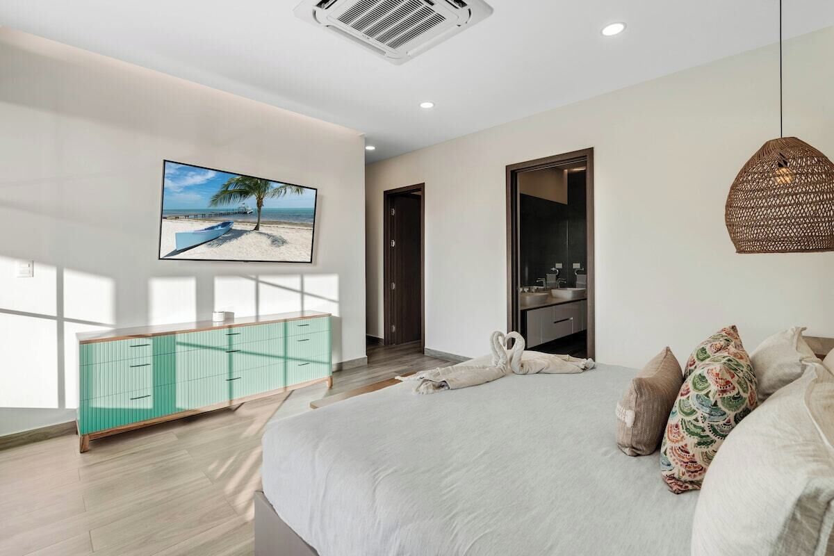 A bedroom with a bed, a television mounted on the wall, a bathroom with a mirror, and a view of the beach.
