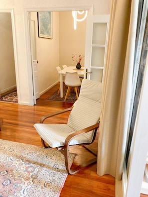 A room with a wooden floor, a patterned rug, a white chair, a dining table with chairs, and a vase with flowers.