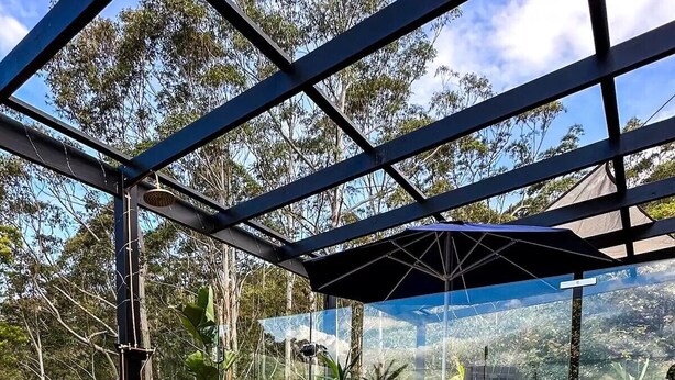 Jacuzzi con cubierta de madera y cerramiento de vidrio, rodeado de abundante vegetación y cielo despejado.
