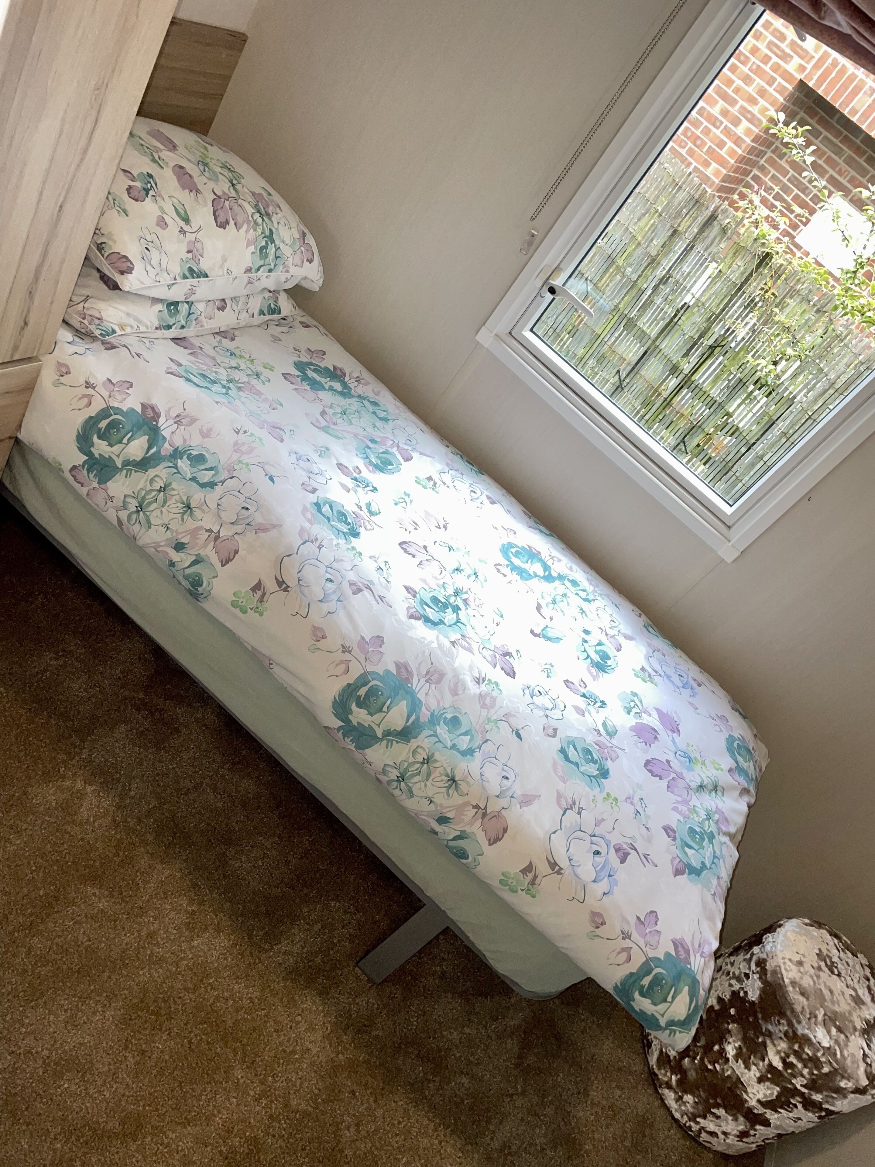 A small, single-bed room with a floral-patterned bedspread, a window with a view of greenery, and a brown carpet.