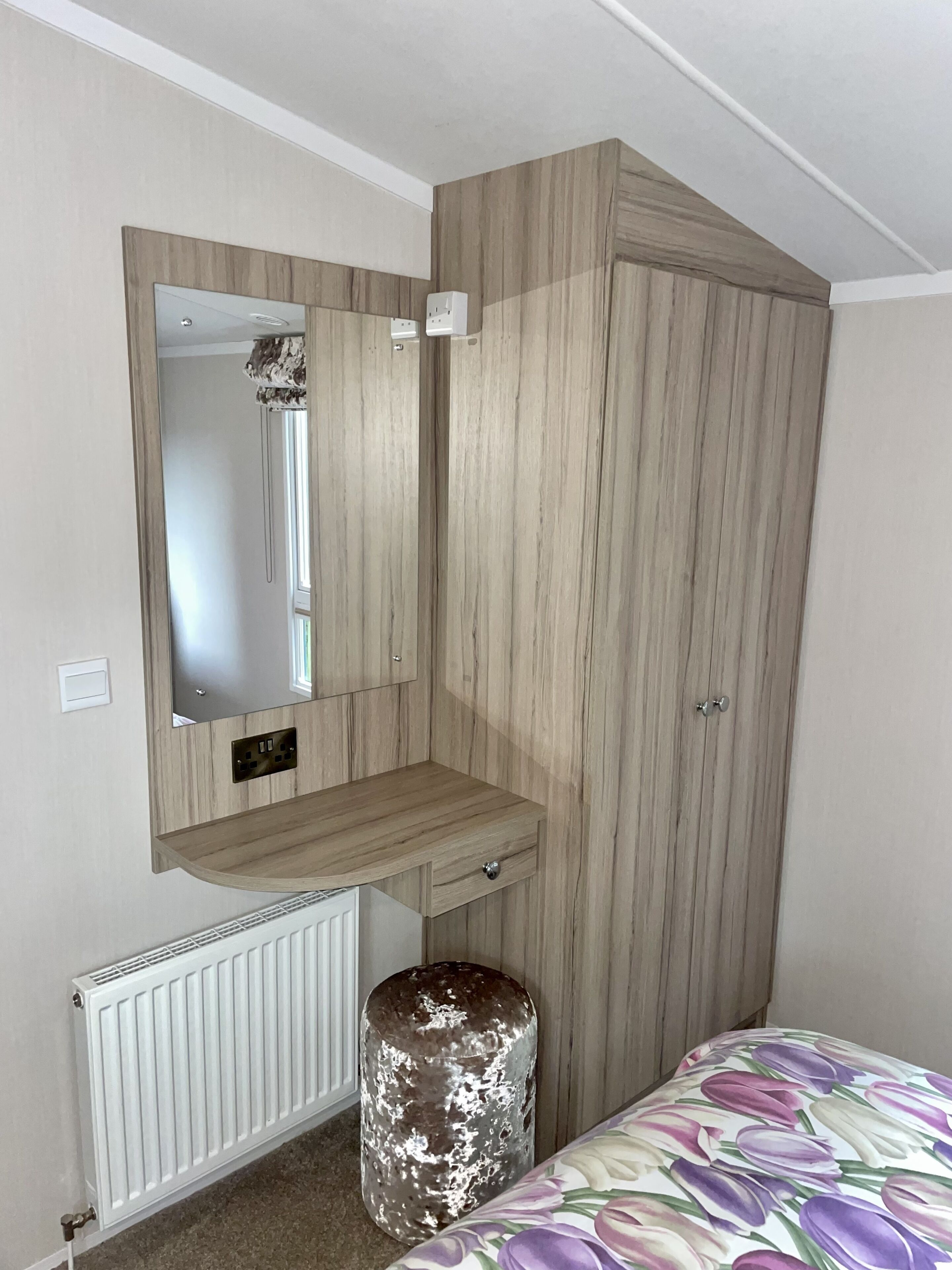 Une chambre avec une armoire en bois, un miroir, une petite étagère, un radiateur et un lit recouvert d’une couvre-lit à motifs floraux.