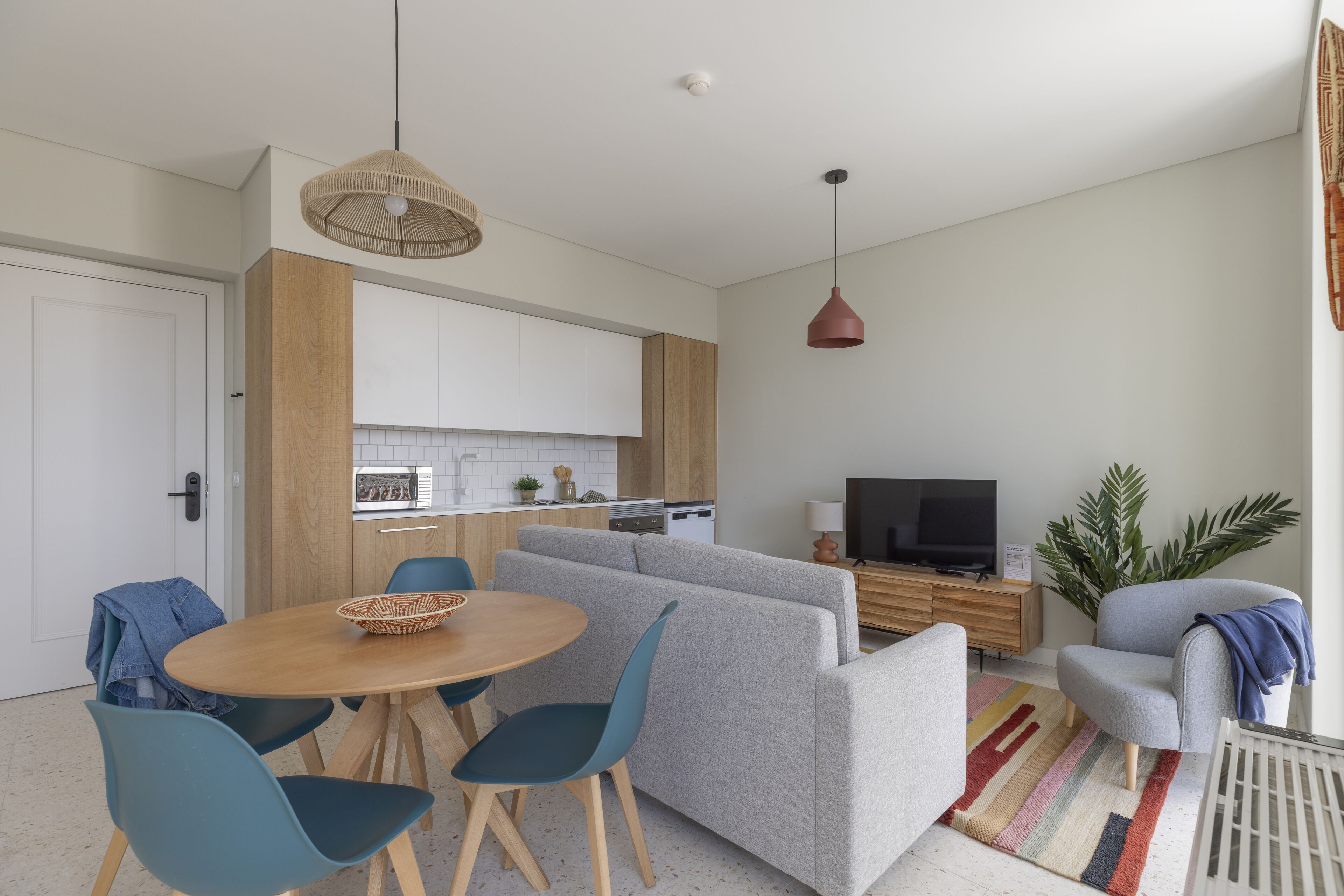 Una sala de estar moderna con un sofá gris, sillas azules, una mesa de comedor de madera y una cocina al fondo.