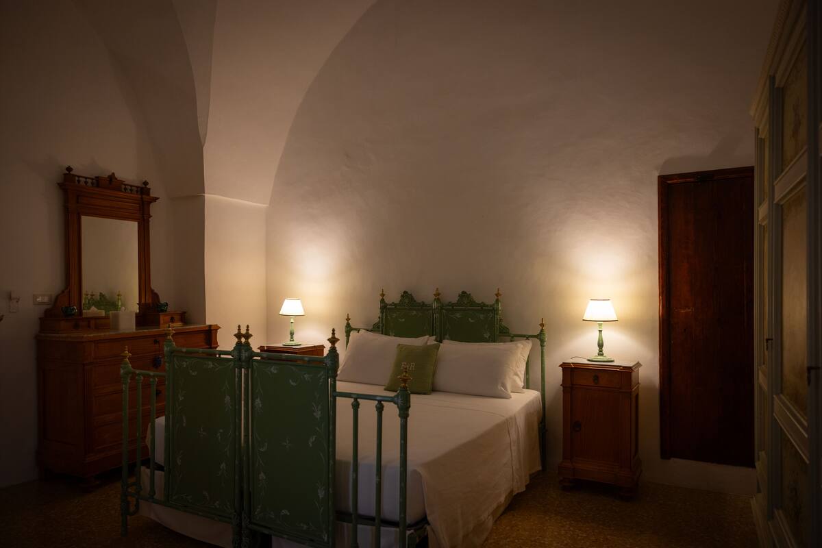 A bedroom with a green bed frame, two bedside tables with lamps, a wooden dresser, and a mirror on the left wall.