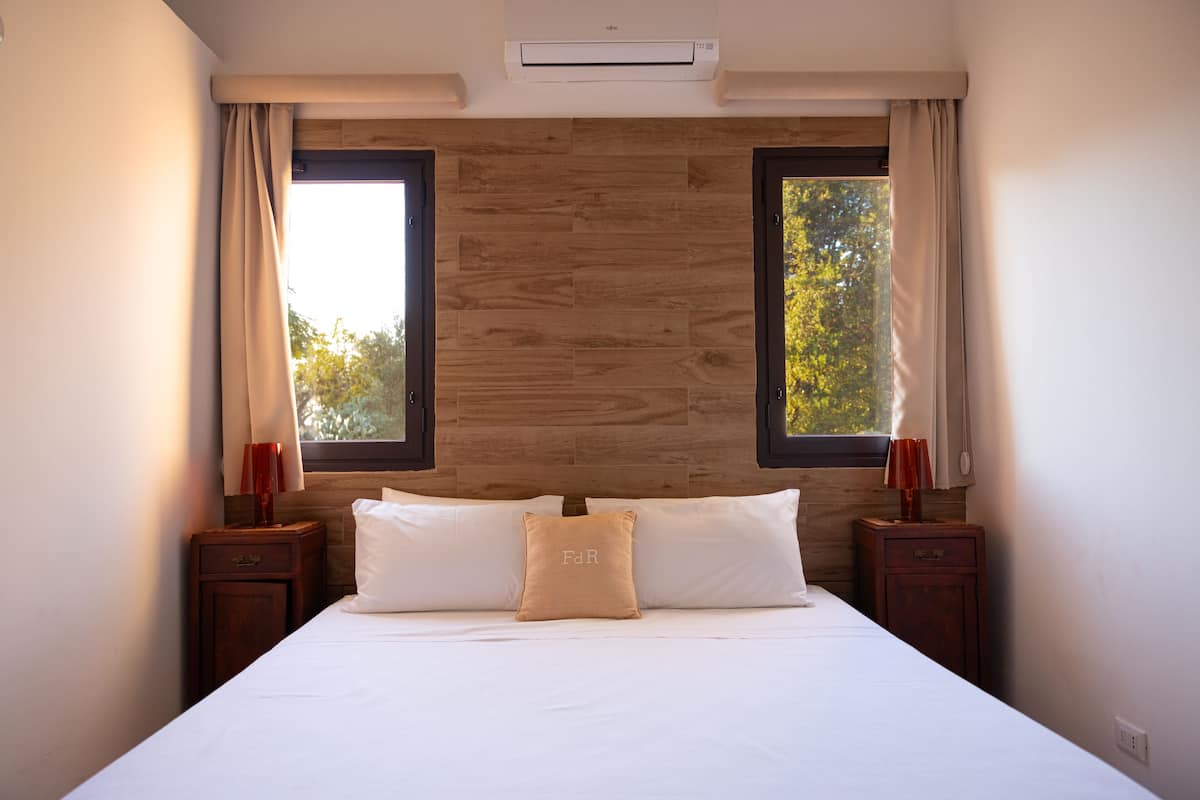 A bedroom with a wooden headboard, two windows with curtains, a bed with white linens, and two bedside tables with lamps.