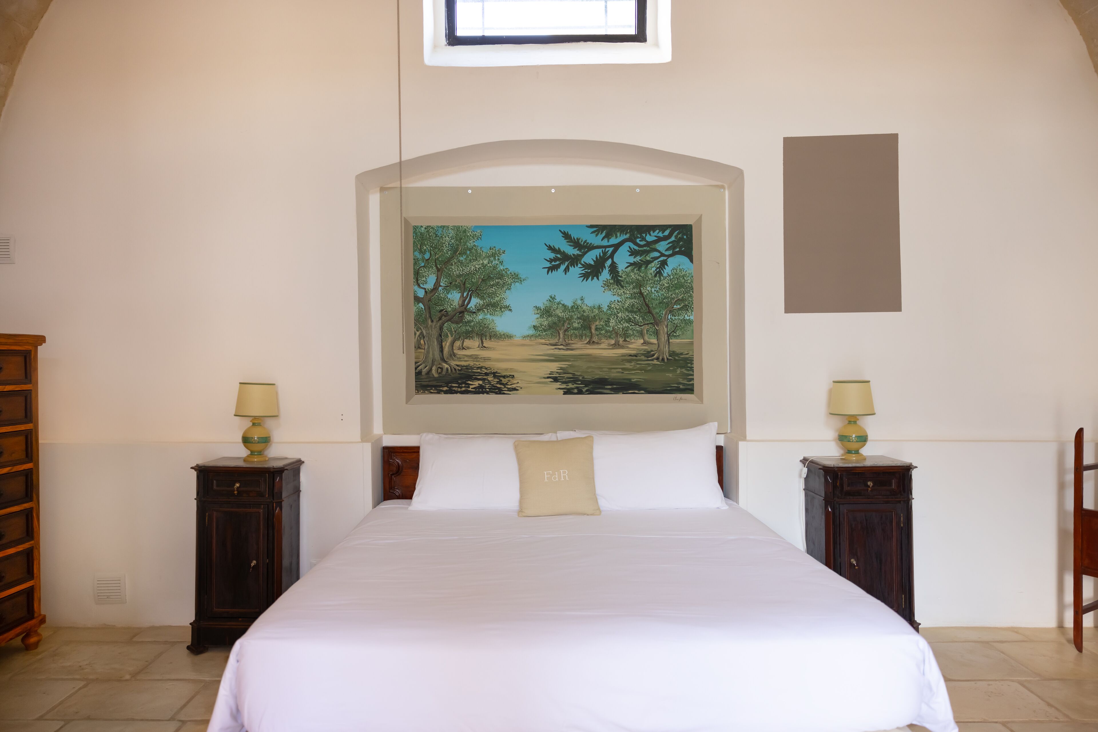 A bedroom with a large bed, two bedside tables with lamps, a painting above the bed, and a wooden chair in the corner.