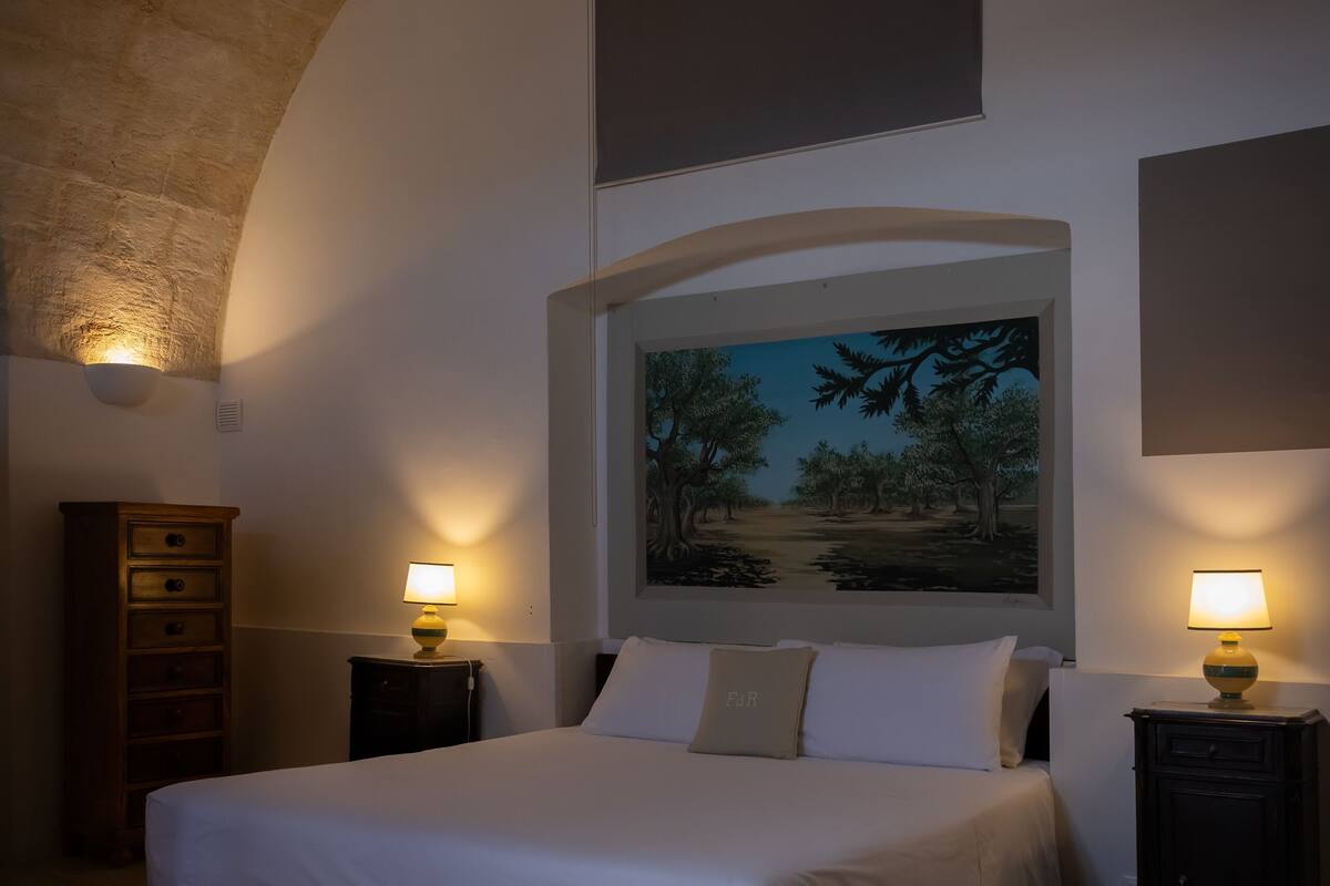 A bedroom with a large bed, two bedside tables with lamps, a wooden dresser, a painting above the bed, and a stone ceiling.