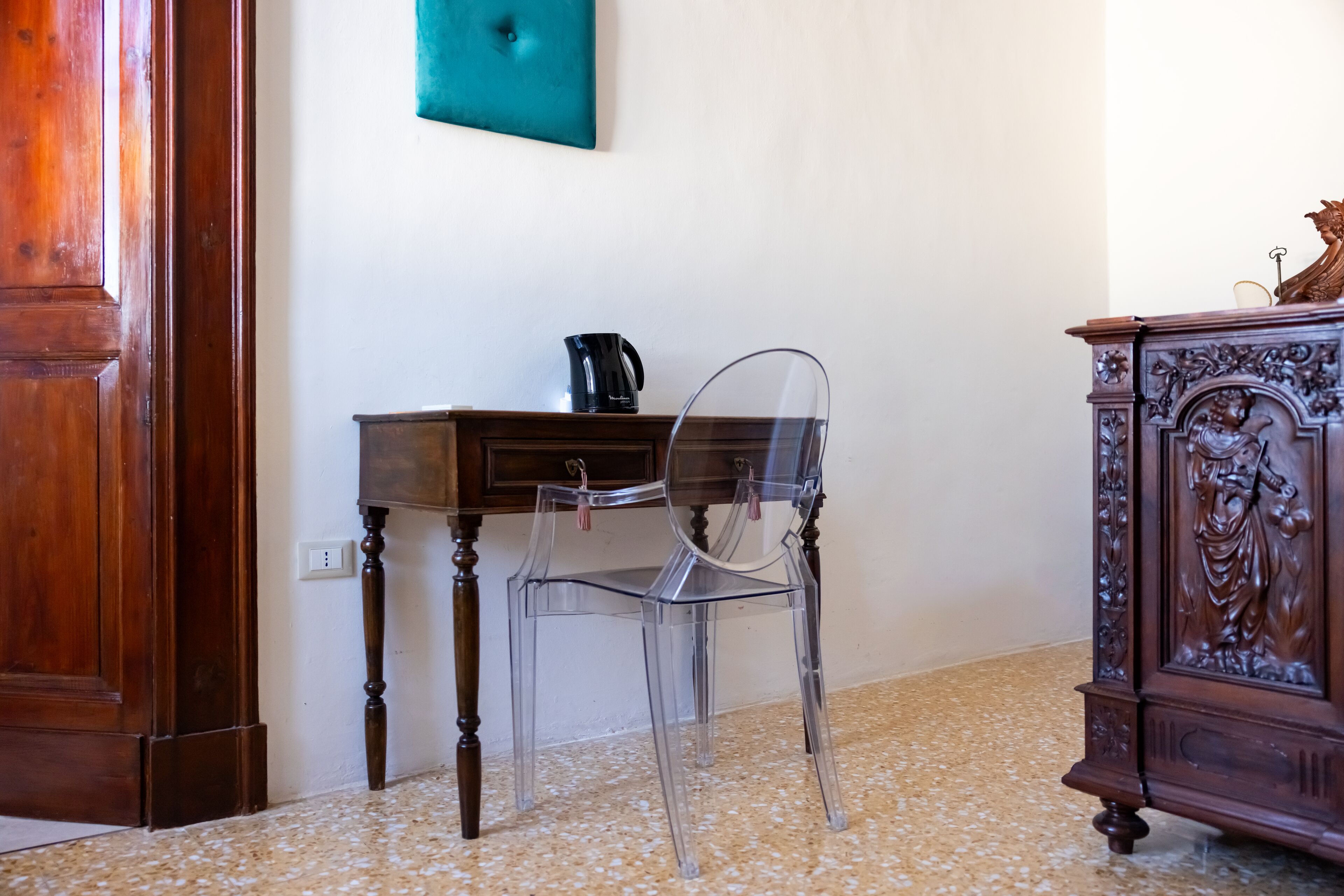 A room with a wooden desk, a clear plastic chair, a dark wooden cabinet, and a green wall decoration.