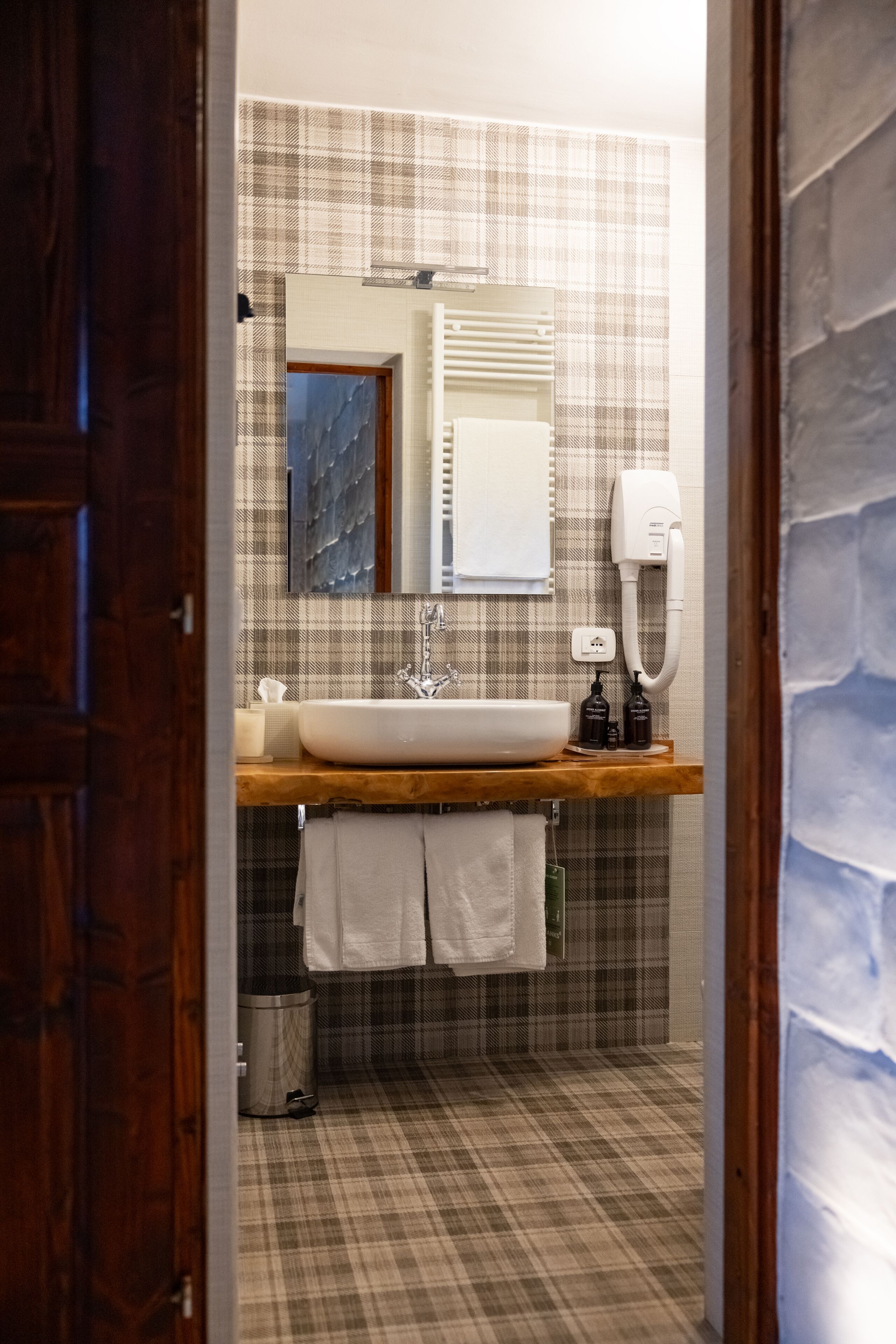 A bathroom with a wooden vanity, a white sink, a mirror, a towel rack, and a trash can.