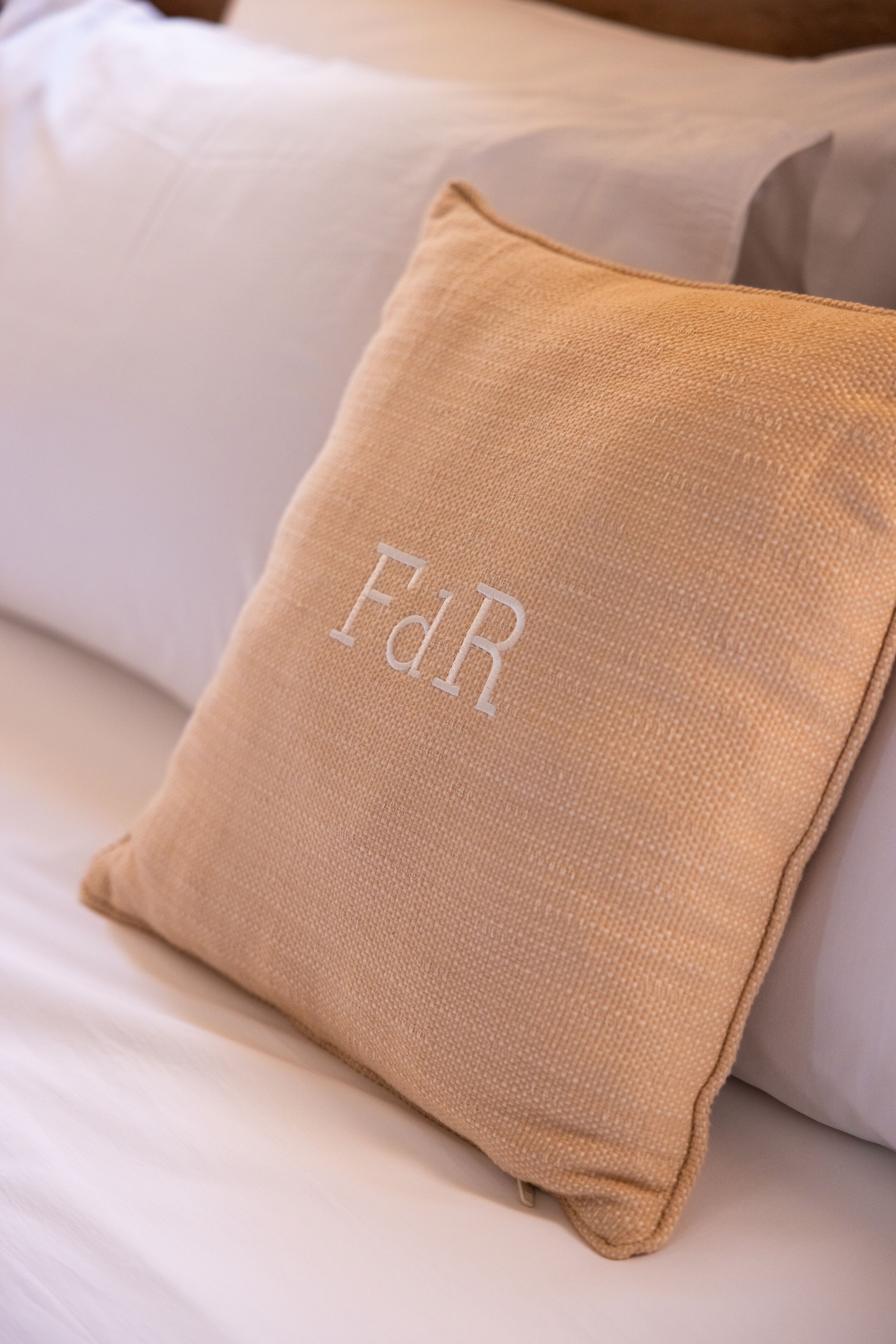 Una cama con ropa de cama blanca y una almohada color beige con iniciales bordadas.