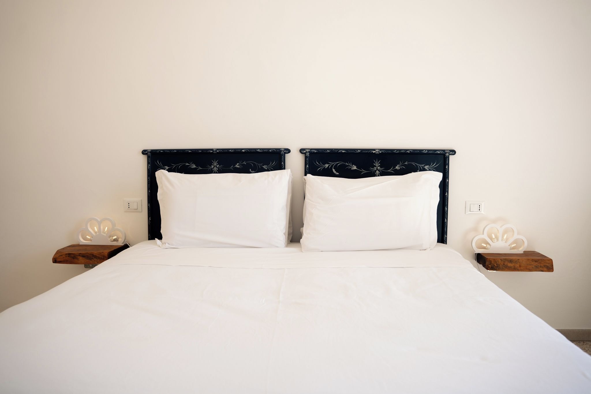 A double bed with a dark headboard and white bedding, flanked by wooden nightstands with white decorative elements.