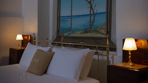 A hotel room with a bed, two bedside tables with lamps, a large window with a painting of a tree and sea view, and a small cabinet.