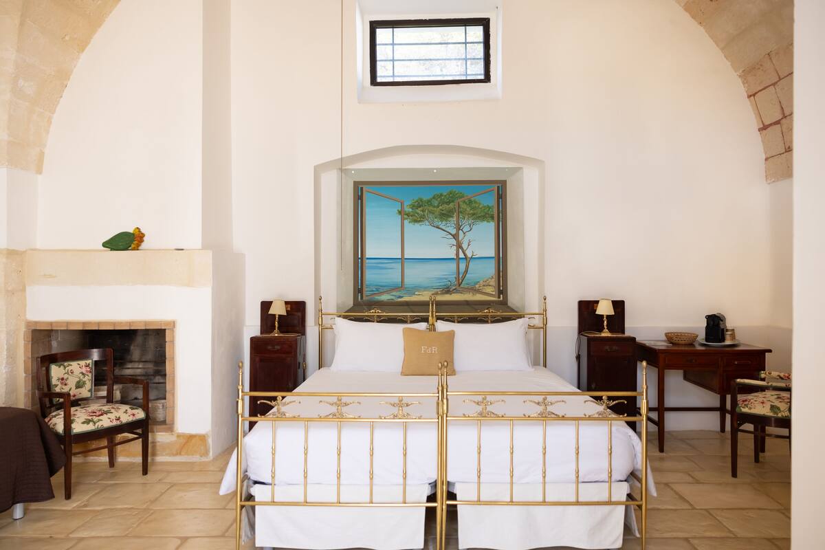 A bedroom with a bed, a painting of a tree by a sea, a fireplace, and a desk with a lamp.