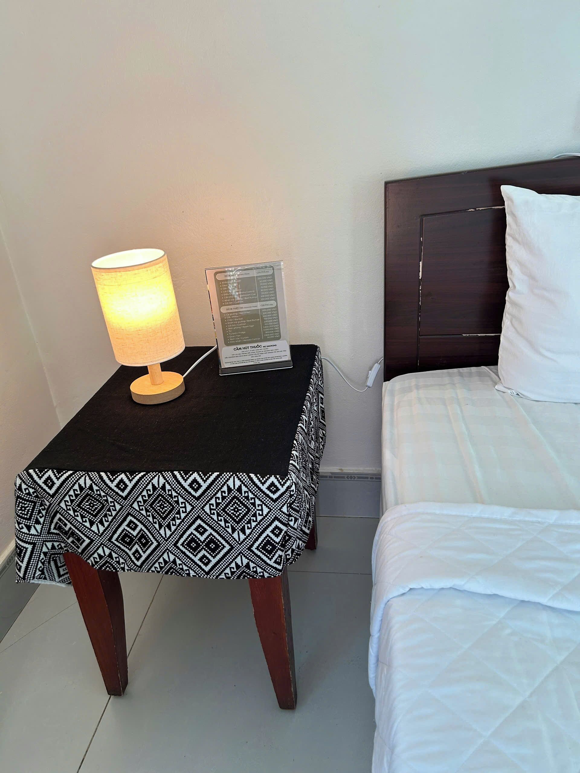 A neatly made bed with a white pillow and a black and white patterned tablecloth on a small wooden nightstand beside it.