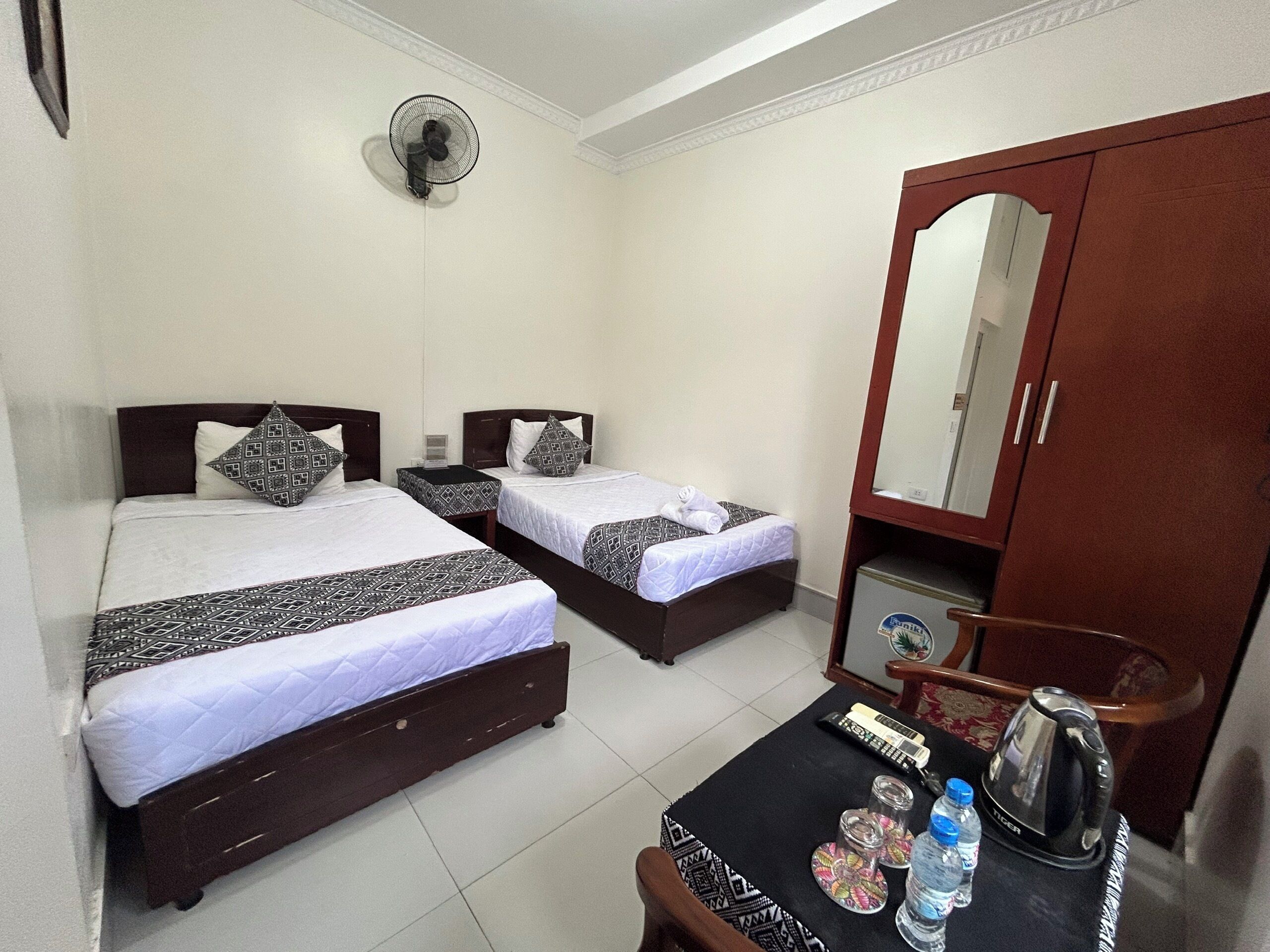 A hotel room with two beds, a table with a kettle, a water bottle, and a mirror.