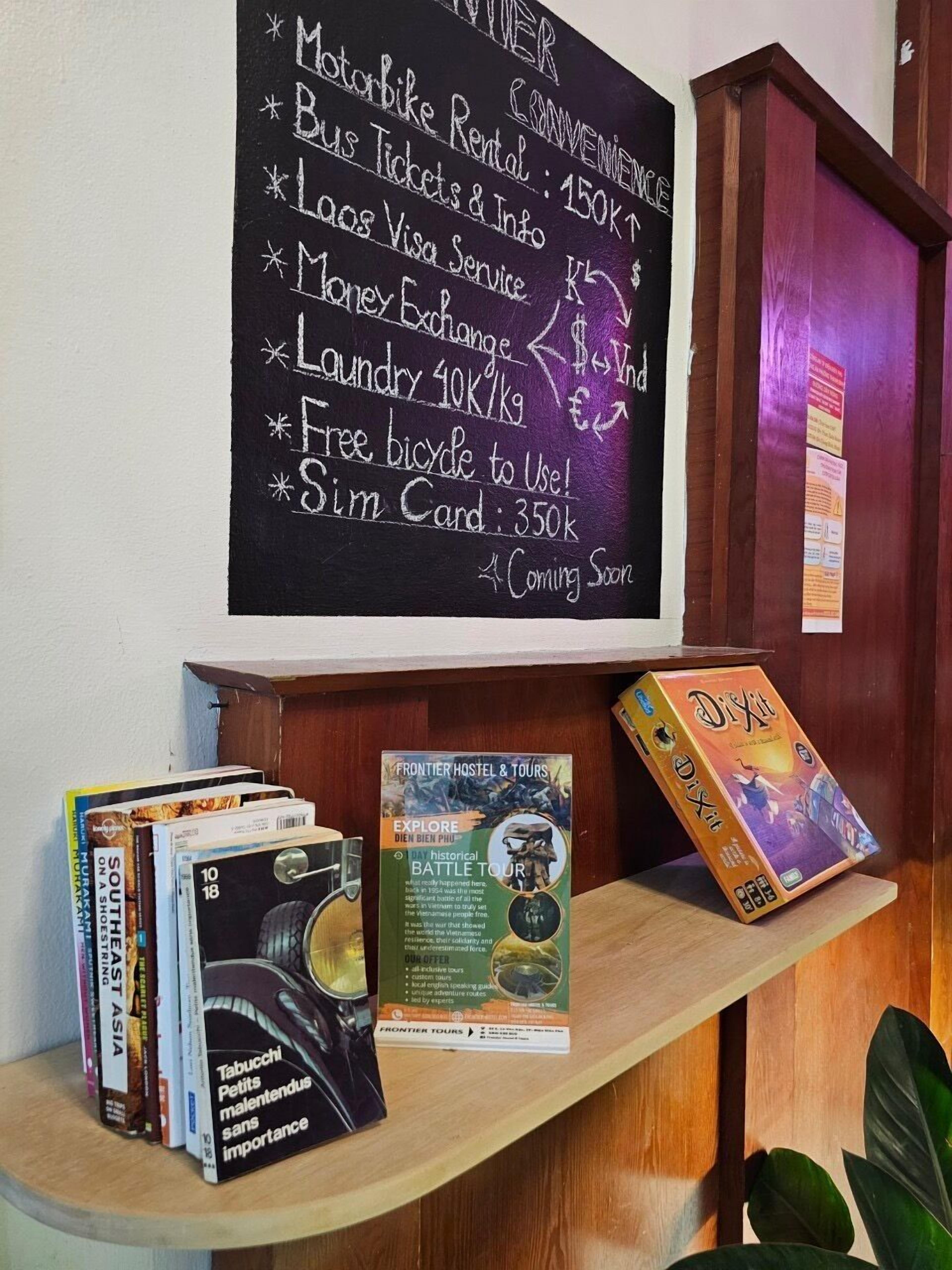 A room with a chalkboard listing amenities like motorbike rental, bus tickets, and a free bicycle. There's a bookshelf with books and a game titled 'Dixit'.