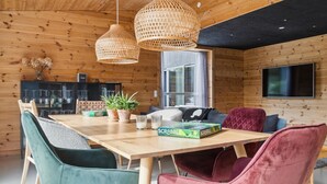 Ein gemütliches Holzhäuschen mit Esstisch, Stühlen, Fernseher und einem Fenster mit Vorhängen.