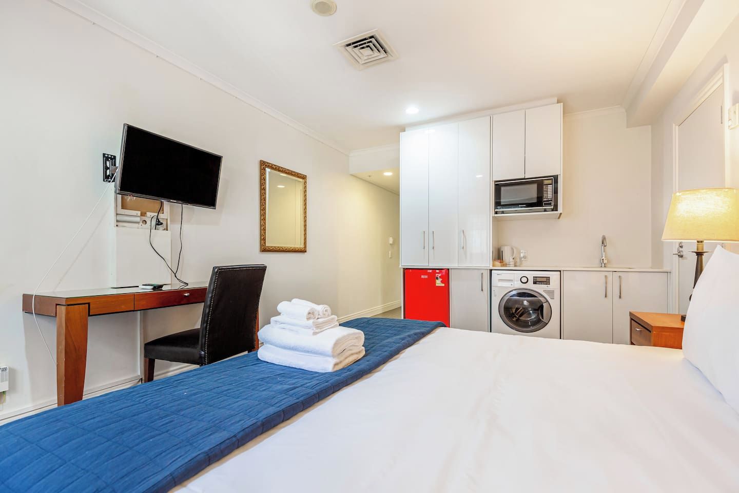 A hotel room with a bed, a desk, a chair, a TV, a mirror, a microwave, a washing machine, and a red cabinet.