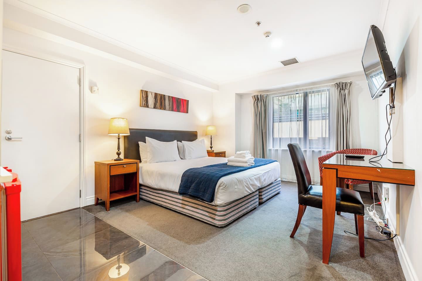 A hotel room with a bed, bedside tables, a desk with a chair, a television, and a window with curtains.