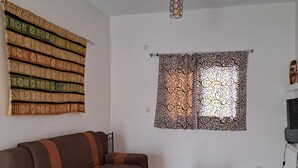 A living room with a brown sofa, a small round table, a decorative wall hanging, and a patterned window treatment. - Villa Neuve, Aérée et Confortable (Nianing)