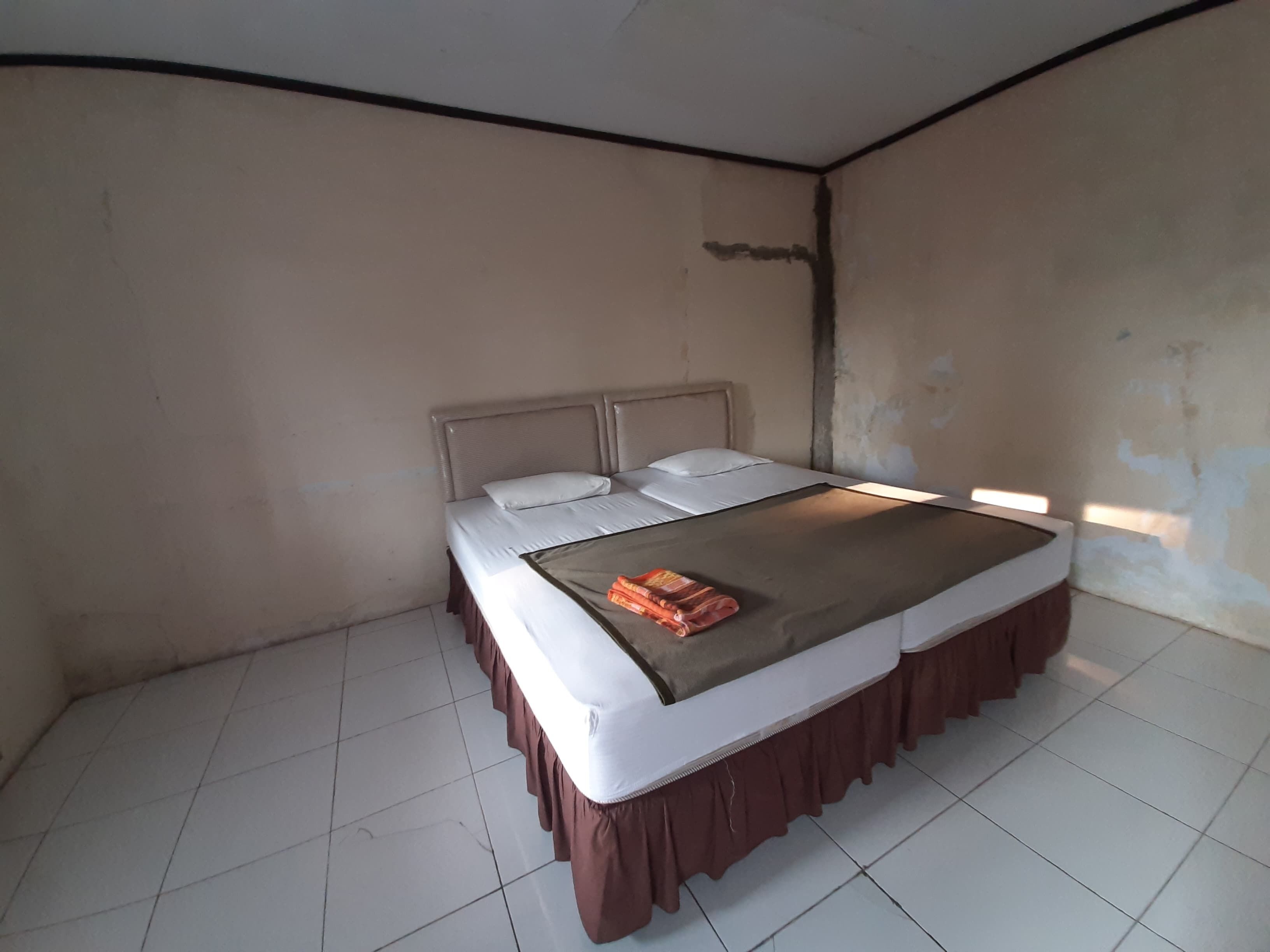 A single bed with a headboard, white bedding, and a brown skirt. The room has plain walls and a tiled floor.