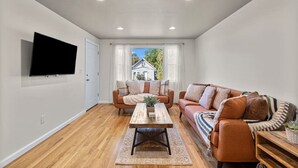 Una sala de estar con sofá, una mesita de centro, un televisor montado en la pared y una ventana con cortinas.