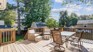 Terraza de madera con mobiliario exterior, incluyendo sillas y una mesa, rodeada de árboles y vistas a casas vecinas.