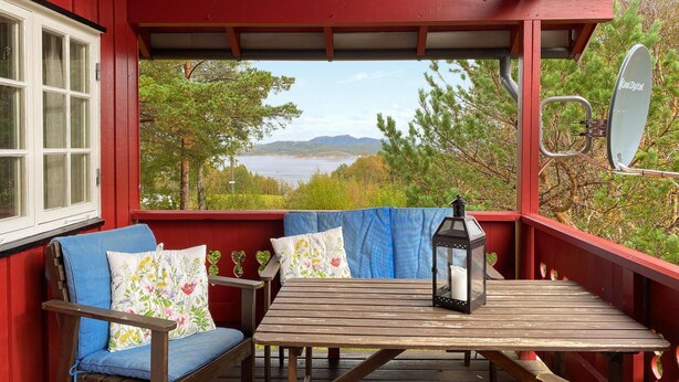 Une véranda couverte avec une table en bois et deux chaises bleues, une lanterne et une vue sur un lac et des montagnes.