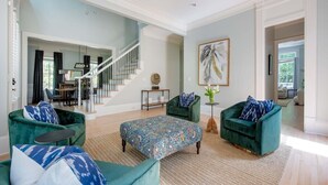Un salon spacieux avec un escalier, des fauteuils en velours vert, un pouf à motifs et un grand tableau abstrait.