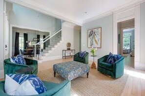 A spacious living room with a staircase, green velvet armchairs, a patterned ottoman, and a large abstract painting.