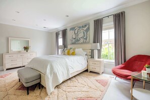 Een slaapkamer met een bed, nachtkastjes, een stoel, een dressoir, een spiegel en een raam met gordijnen.