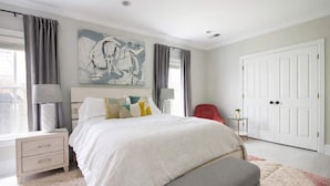 A bedroom with a large bed, a bedside table, a lamp, a chair, a small table with a plant, and a door.