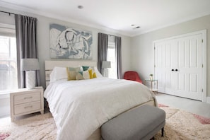 Une chambre à coucher avec un grand lit, une table de chevet, une lampe, une chaise, une petite table avec une plante et une porte.