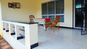 A room with a tiled floor, a table with a patterned tablecloth, two wicker chairs, a bench, and a window with metal bars. - Rena Home Stay (Yercaud)