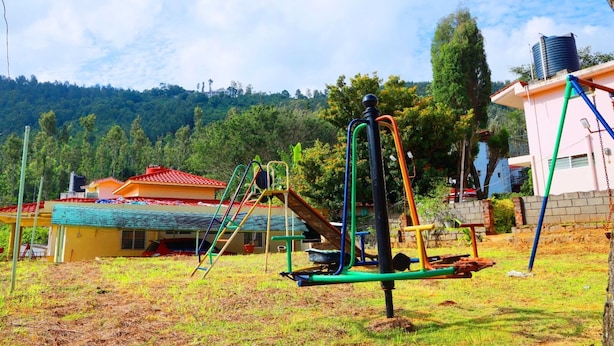 A playground with various equipment, a building with a red roof, and a water tank in the background. - Rena Home Stay (Yercaud)