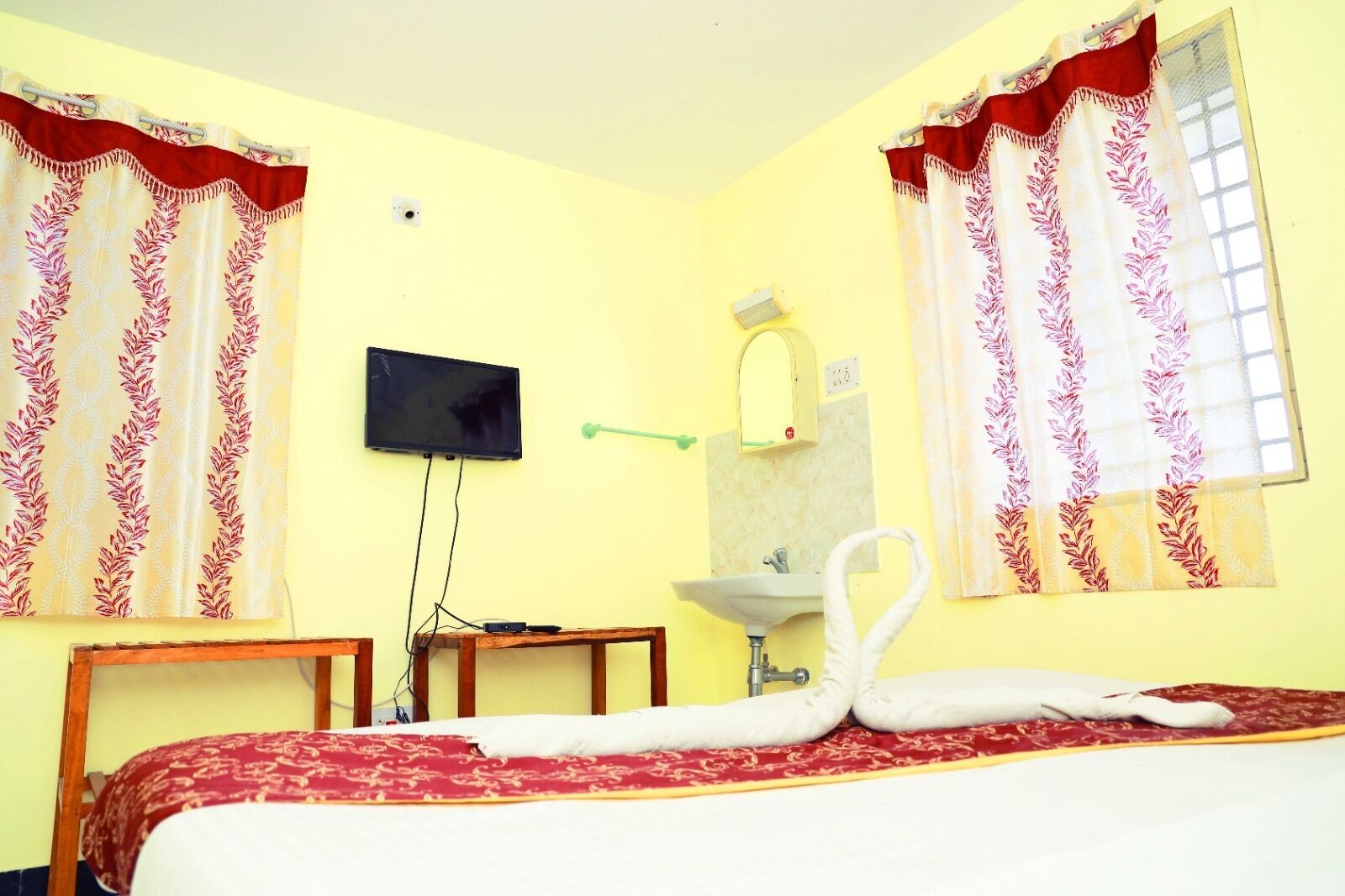 A hotel room with a bed, a television mounted on the wall, a sink, and two windows with curtains.
