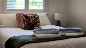 Un lit bien fait, avec des oreillers, une couverture et des serviettes pliées. Il y a une fenêtre avec des stores, une lampe et une vue sur la verdure à l’extérieur.