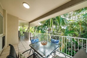 Un balcon doté d’une table et de chaises en verre, entouré d’une balustrade blanche, et offrant une vue sur un jardin luxuriant planté de palmiers.