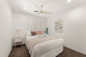 Ein Schlafzimmer mit einem Bett, einem Nachttisch, einem Deckenventilator, einem Fenster mit Jalousien und einem gerahmten Bild an der Wand.