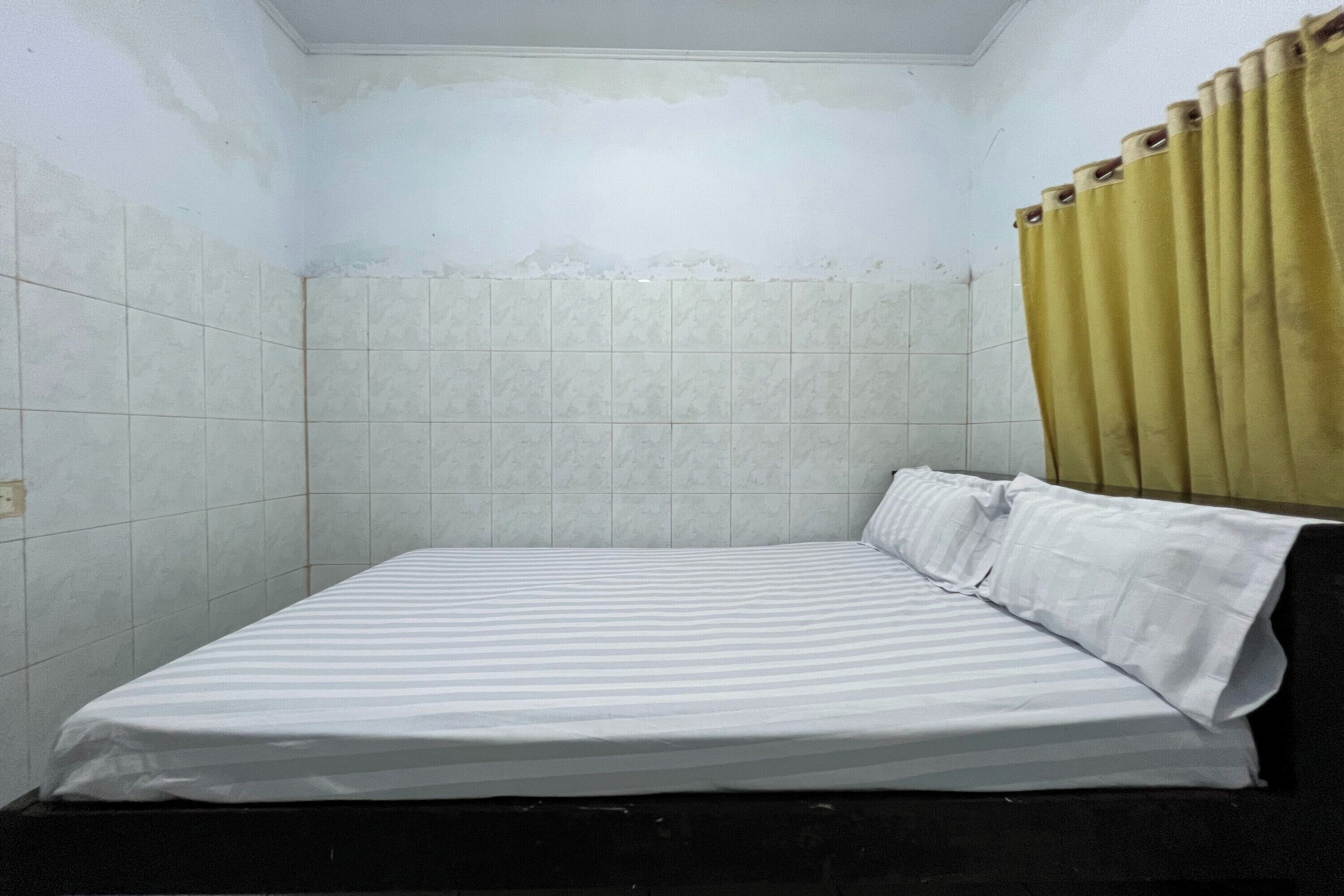 Une petite pièce étroite, avec un lit simple, des murs unis et un rideau.