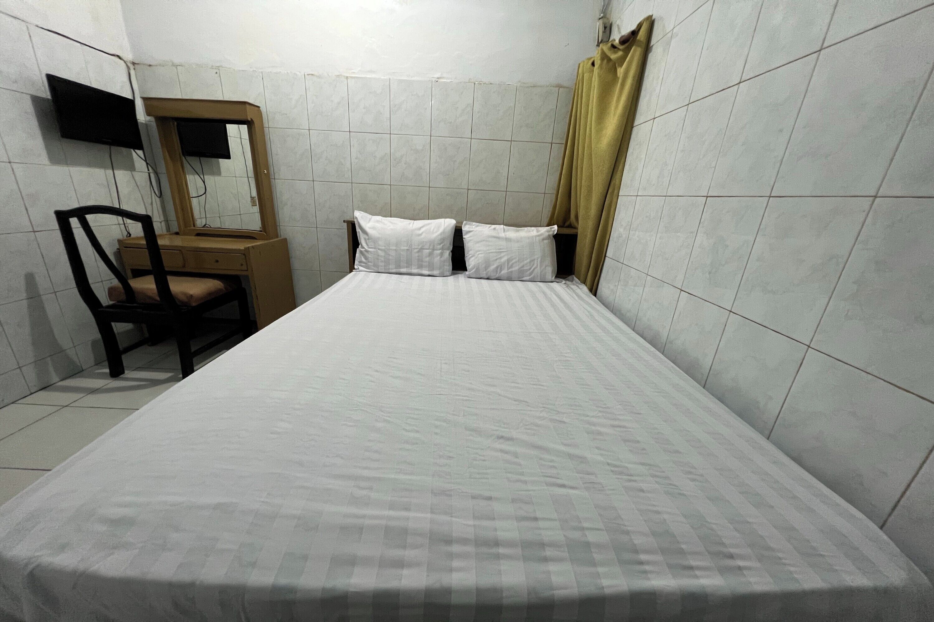A single bed with a white mattress and two pillows, a wooden chair, a small wooden dresser, a mirror, and a television mounted on the wall.