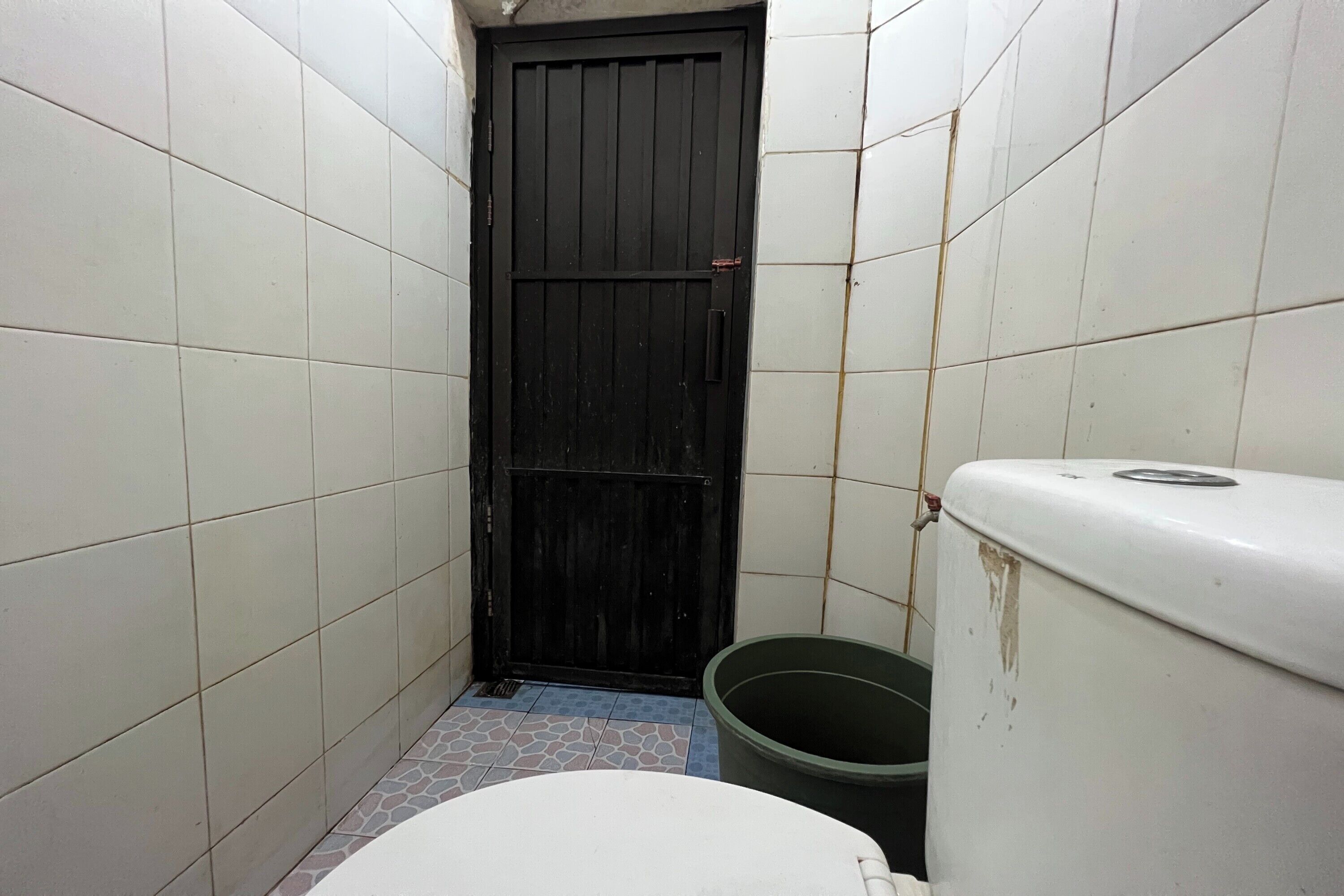 A small, narrow bathroom with white tiled walls, a black door, a white toilet, and a green bucket.