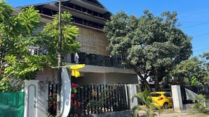 Un immeuble d’habitation doté d’un balcon, une voiture jaune garée devant, et un grand arbre.